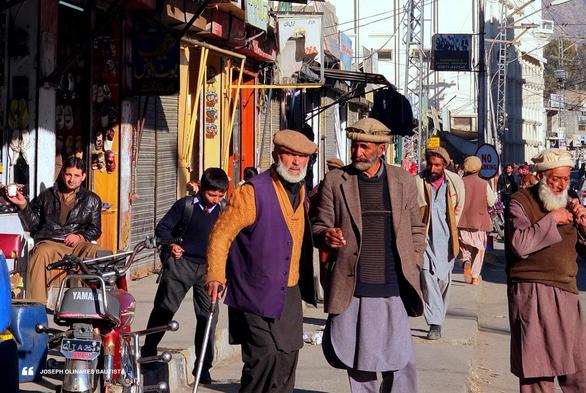 A street scene in the mountain town of Gilgit in Pakistan, with men in traditional attire walking along a pavement lined with small shops. An elderly man in a purple vest and cap leans on a cane, accompanied by another elderly in a coat and a hat, while a younger man in a leather jacket sits nearby. Shop fronts display colorful signs and goods, with the mountain faintly visible in the background. Photo taken by Joseph Olinares Bautista in Gilgit, Pakistan. 2013.