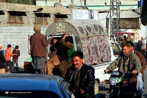In a bustling street of northern Pakistan, a small van with faded floral designs and patterned curtains is packed with men, one of whom stands at the open back, gripping the frame and peering into the sunlit road ahead. Motorcyclists weave past, faces weathered but alert, wrapped in winter layers. Behind them, children linger near a wall, sharing a quiet moment as sunlight spills across the rough stone buildings and painted shopfronts. The cold November air feels alive with movement and routine. Photo taken by Joseph Olinares Bautista in Gilgit, Pakistan. 2013.