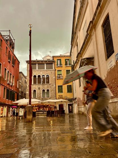A rainy Venetian square with glistening wet stone pavement. People with umbrellas hurry across the scene, their movements slightly blurred. In the background, colorful historic buildings - red, yellow, and brick - frame a small café with white parasols. 
The sky is heavy and grey-green, adding a dramatic mood to the scene. A tall red flagpole stands near the center, enhancing the vertical rhythm of the square.