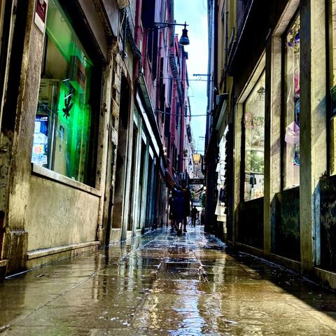 A narrow Venetian alley after rain, its wet stone pavement reflecting shop lights and the blue sky. On the left, a green-lit ATM glows; on the right, shop windows display clocks and colorful items. The buildings are tall, close, and slightly leaning inward. Two people walk in the distance, framed by the tight perspective of the alley. The mood is moody yet vibrant, with a sense of quiet after the storm.