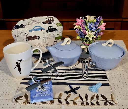 Tea tableau celebrating Father's Day with a classic car themed tea cozy, a mug with a silhouette of a boy flying a model airplane over his head, vintage Homer Laughlin cream and sugar set, paper flowers, a dynamic sculpture of flying airplanes, glass miniatures of a jet and a space shuttle, and Double Bergamot Earl Grey tea from Stash.