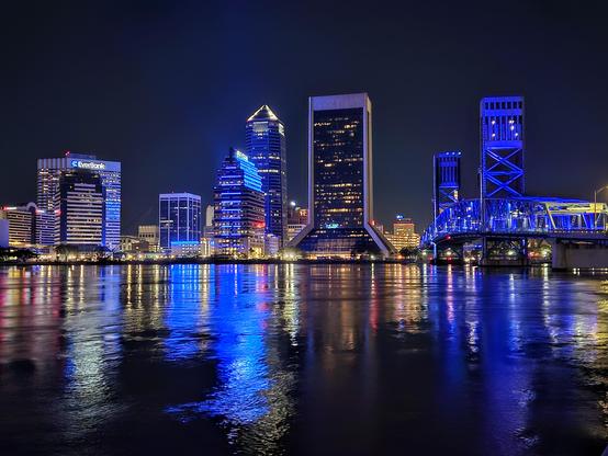 Late night view across the wide, dark river where the downtown skyline and iconic Main Street Bridge glow from blue night lighting and reflect onto the water below.
