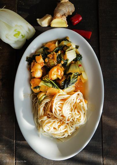 Dunkler Holzuntergrund. Ovaler weißer Teller. Darin: Geschmorter Pak Choi mit Bananen, Ingwer, Peperoni, Zwiebeln, Kreuzkümmel und mit Ramen-Nudeln