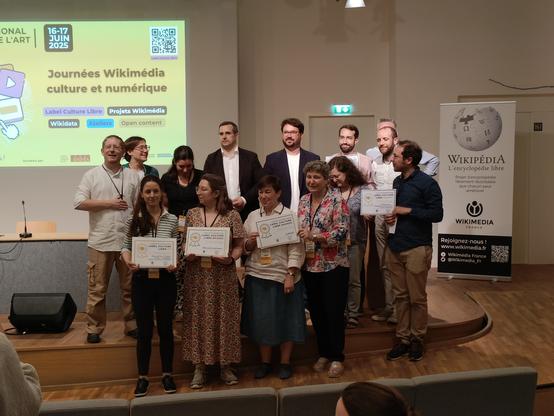 Photographie numérique couleur en format paysage.
Photographie de groupe du jury et des labellisés, qui tiennent leur diplôme. Un écran à gauche et un kakémono Wikimedia France à droite.