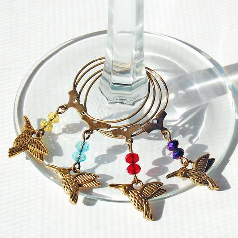 Set of four gold-tone hummingbird wine glass charms with colored beads in yellow, blue, red, and purple, displayed around the base of a clear wine glass.