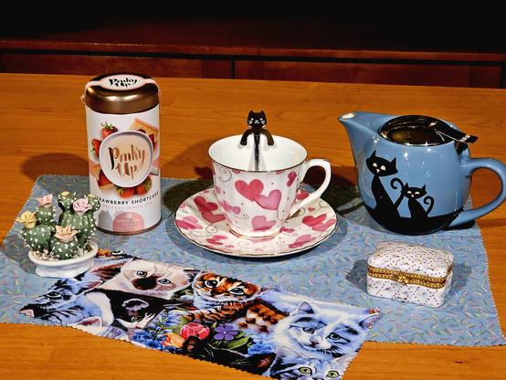 Tea tableau with a cat theme. On a blue tea towel are a blue Cattitude teapot, white cup and saucer decorated with large pink hearts and with a cat shaped spoon standing in the cup, a panel of kitten patterned fabric, a painted bisque sculpture of flowering cactus, a small white and pink trinket box, and Pinky Up Strawberry Shortcake black tea.