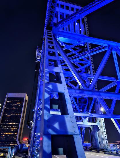 From pedestrian crossing, late night, looking upwards into the huge metal bridge structure, glowing from blue night lighting.