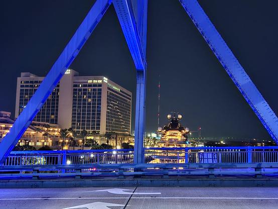 From pedestrian crossing, late night, looking upwards into the huge metal bridge structure, glowing from blue night lighting.