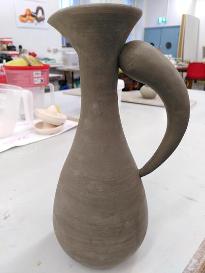 Small ceramic water drop pitcher at the greenware stage. The body is a water drop shape and the handle is an upside down arched elongated drop.