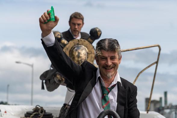 A cheerful man in a suit and green striped tie, with a playful moustache and goatee, smiles at the camera while holding up a small green plastic water pistol. Behind him, another man in a suit stands with a large, decorative shield, both on a boat during The Family Shenanigans parade in Cork.