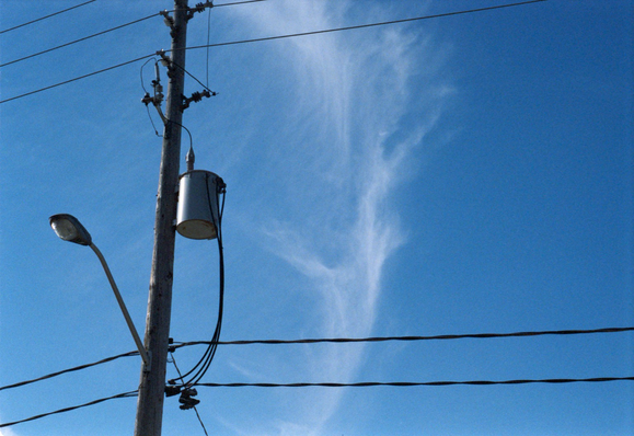 A wisp of cloud across a blue sky behind a wooden utility pole festooned with transmission lines, insulators, a transformer bucket, and a street lamp.
