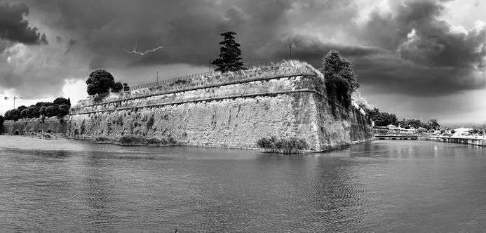 Ein Panoramablick in Schwarz-Weiß auf eine steinerne Festungsmauer an einem ruhigen Gewässer, mit dramatischen dunklen Wolken über dem Kopf und einem Blitz am Himmel. Die Szene vermittelt ein Gefühl von Vorahnung und historischer Architektur inmitten einer natürlichen Landschaft.