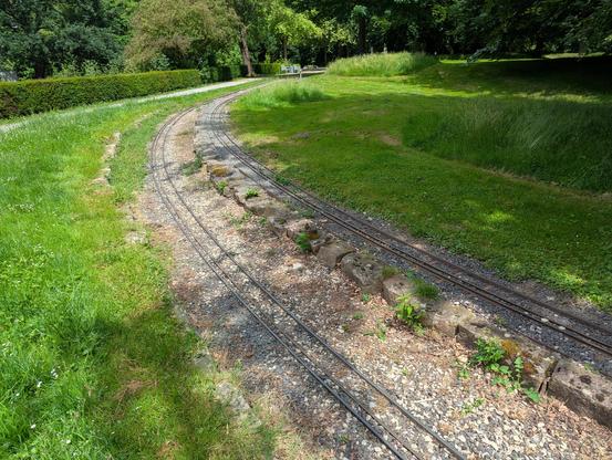 Two parallel model train tracks put on loose gravel. They are surrounded by green grass. Both tracks have a third rail, for variable gauge (~18.4 cm and 12.7 cm).  To the left, there is a narrow gravel trail, leading through the park.