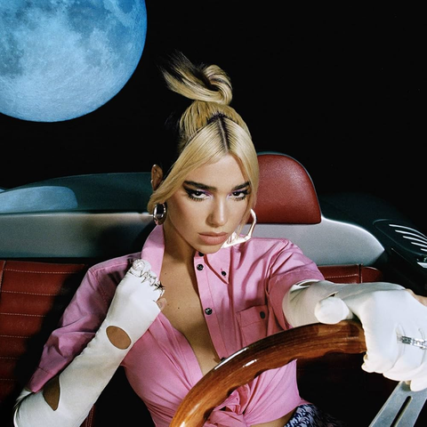 Dua Lipa's Future Nostalgia album cover. A woman dressed in a pink shirt that is open exposing a portion of her chest and long, white gloves. She is sitting looking downward in the back of a red and white car with her hand on the steering wheel. The background dis all black with the moon looking huge right behind her on the left.