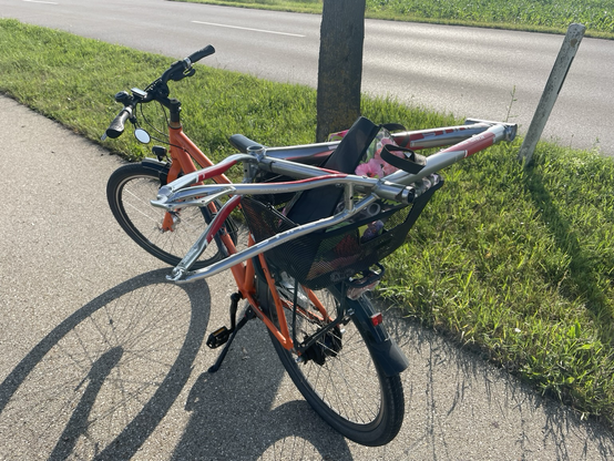 Ein oranges Fahrrad steht auf dem Radweg, auf dem Gepäckträger ist ein Korb, darauf ein Fahrradrahmen ihn irgendein Anbauteil.