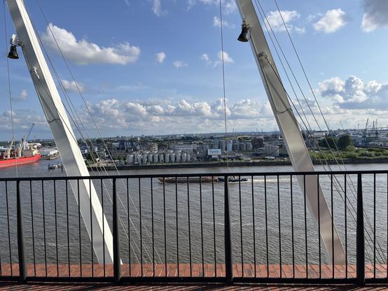 A view of a river with a red ship and industrial buildings along the shoreline. A metal railing is visible in the foreground, with a blue sky and scattered clouds overhead.