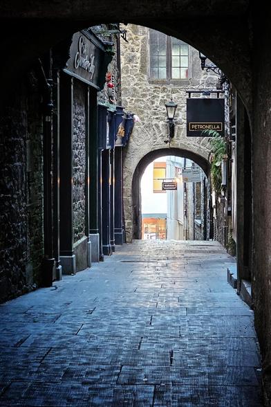 A narrow, dimly lit cobblestone alleyway with stone buildings on either side, leading through a stone archway towards a brighter, open area. Various shop signs, including "Sweater Shop" and "PETRONELLA," are visible on the buildings. A street lamp hangs above the arch, illuminating the path. The overall mood is historic and inviting.