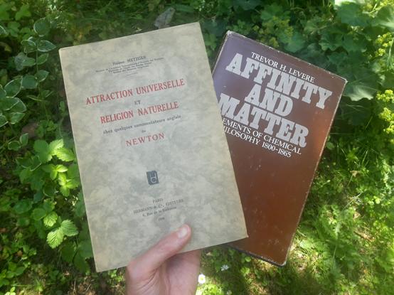 A hand holds up two books. One is called "Attraction universelle et religion naturelle chez quelques comentateurs anglais de Newton", by Helene Metzger, from 1938. The other one is called "Affinity and Matter, elements of chemical philosophy 1800-1865", by Trevor H. Levere.