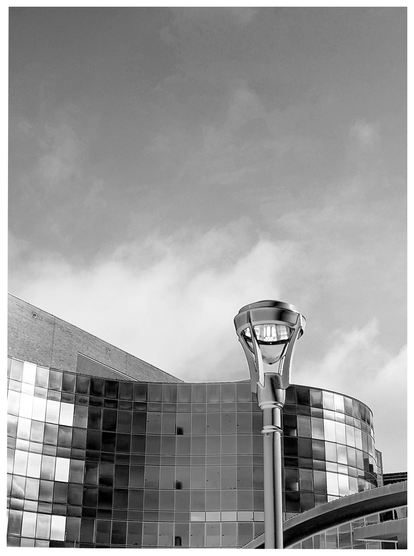 AI-assisted alt text:

	
in the distance,  a modern glass building with reflective windows curves out from a brick facade. in the  foreground, top top of a street lamp appears same size as the building. The sky above is partly cloudy
