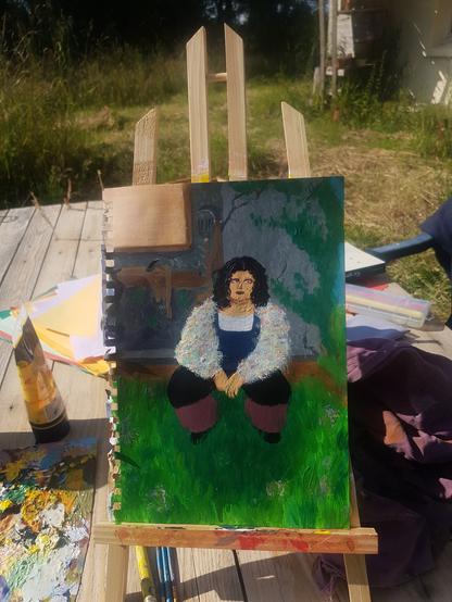 Photo d'une peinture à l'acrylique sur un chevalet. Est représenté une personne, la peau blanche, les cheveux noirs et bouclés, les yeux bleus et du far à paupière et rouge à lèvres bordeaux.
Iel est accroupie dans l'herbe devant un atelier de bricolage dans une maison en ciment grise.