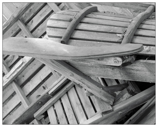 Black and white photograph of a pile of wooden chairs stacked haphazardly. The visible chair on top features curved back slats and armrests, with weathered and chipped wood textures prominently displayed. The underlying chairs are mostly obscured, showing only portions of their wooden slats.