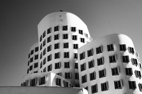 Black and white photo of a modern, curving building with irregularly placed rectangular windows, showcasing a deconstructivist architectural style against a clear sky. The undulating façade creates a sculptural, wave-like appearance.