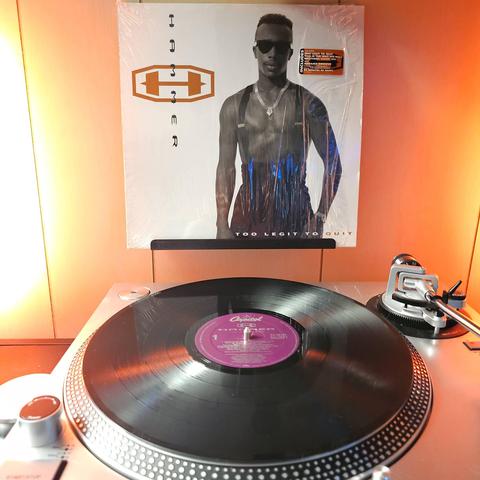 A vinyl record sits on a turntable. Behind the turntable, a vinyl album outer sleeve is displayed. The front cover shows Hammer shirtless wearing suspender pants. a gold necklace, and sunglasses.