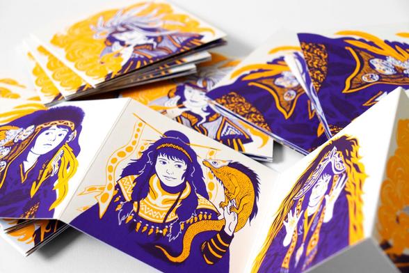 Like in the previous photos, a stack of closed leporellos appears in the background. In this photo, the foreground shows the back side of the leporello. On it, there are four portraits of different women, one on each panel of the accordion fold. Still printed in two colors, yellow and purple, each woman wears an imaginary costume.
