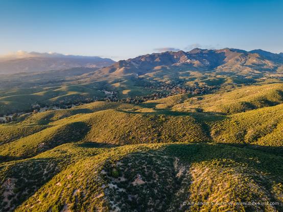 A landscape photo with an elevated view across rolling hills covered with low-growing shrubby vegetation and a mountain range rising from those hills towards a clear sky. The scene is illuminated by warm evening light.