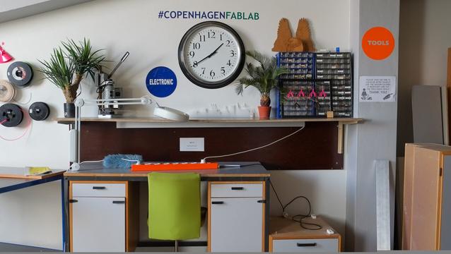 Photograph of one wall of FabLab Copenhagen near the entrance. There is a large clock on the wall and the words (hashtag) FABLAB COPENHAGEN on the wall above the clock. Against the wall is a desk and chair as a soldering station. There is a desk lamp with a magnifying glass on the desk and an electric extension cord. There is a shelf above the desk on which there is a components cabinet, a measuring device, plants and various prototypes. A round vinyl sticker reading ELECTRONICS is on the wall above the shelf and another one reading TOOLS on a column next to the desk. To the left of the desk on the wall there are several reels of 3D printing filament hanging and there are other work tables and cabinets visible. FabLab Copenhagen, Visby, Denmark, 27 June 2018. Photo Cindy Kohtala