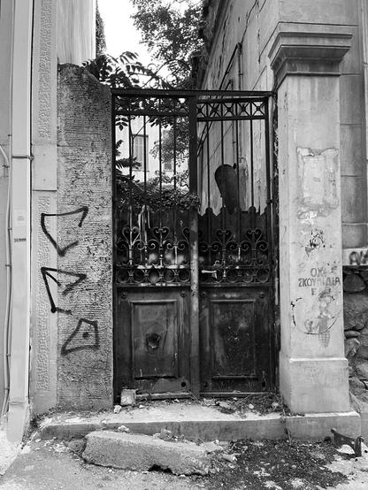 Black and White photo. The gate of a ruined neoclassical house, a typical tall late 19th century garden gate. The supporting column on one side has had its stone facing removed, on the other clumn No Garbage Here has been stenciled. Rubble, graffiti, a glimpse of leaves and a crumbling building through the gate.