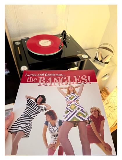 Auf einem schwarzen Plattenspieler läuft eine schwarze Schallplatte. Davor halte ich das Cover ins Bild. Die vier Musikerinnen der Band posieren in kurzen 70er Jahren Kleidern. Oben steht in geschwungener Schrift Titel des Albums.
