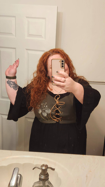 Me, a short fat woman with long red curly hair is posing in front of a mirror with one hand up in the hair and the other holding a phone which obscures my face, although you can tell I'm smiling.

I am wearing a long black chemise with drapey sleeves and over it is an olive green lace up bodice that is cropped to my waist. On the one side are three dandelion fluffs with some of the seeds scattered up towards the shoulder.