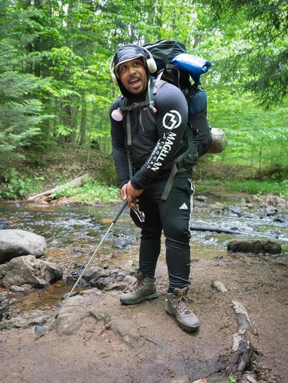 A man next to a stream carrying a camping backpack demonstrates a golf swing using a club that he uses as a hiking pole.