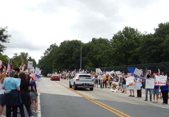 The same crowd of anti-Trump protestors hold signs and flags as they wave to passing cars.