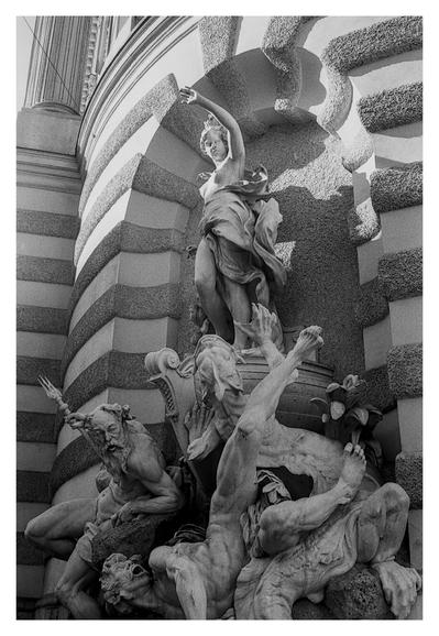 Black and white photograph of “Die Macht zur See”, a monumental wall fountain on the façade of the Michaelertrakt of the Hofburg on Michaelerplatz in Vienna's 1st district Innere Stadt. The fountain was built from 1893 by the sculptor Rudolf Weyr, unveiled in 1895 and is a listed building. The Macht zu Lande fountain is located on the opposite side of Michaelerplatz. The main figure is an allegorical representation of Austria standing on the bow of a ship. A rock architecture with a depiction of the sea god Neptune, a fallen giant and a sea monster rises above the fountain basin. Source of the fountain description: Wikipedia

Translated with DeepL.com (free version)