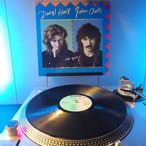 A vinyl record sits on a turntable. Behind the turntable, a vinyl album outer sleeve is displayed. The front cover shows portraits of Daryl Hall and John Oates.