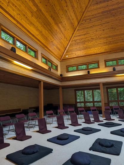 Interior of the Omega Center sanctuary, with warm wooden walls and pillars, as will a vaulted ceiling with small windows near the top of the walls, and large windows against the rear wall, n front of a set of dark-colored mats and cushions for meditation on a beige carpet.