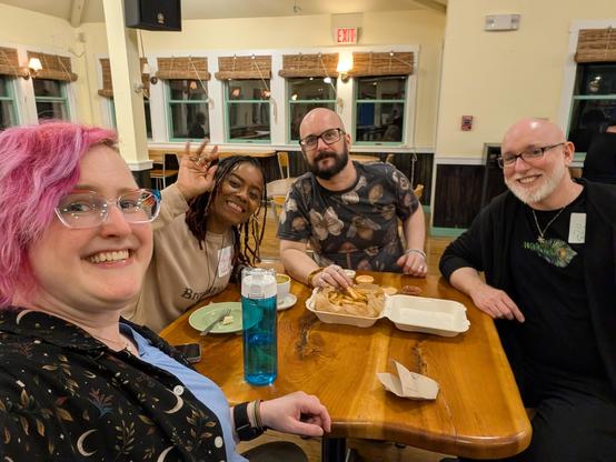Enfys Book, Aly Kravetz, Mat Auryn, and Storm Faerywolf sitting at a cafe table, smiling and waving at the camera.