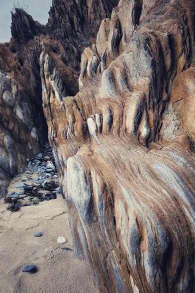 A close-up, low-angle shot of a towering, intricately layered rock formation on a sandy beach. The rock exhibits striking horizontal and undulating patterns in shades of brown, grey, and ochre, with a rugged, textured surface. To the left, a small collection of smooth, grey and white pebbles are scattered on the light brown sand, leading into a narrow crevice between the rocks. The sky above is overcast, and subtle shades of purple and blue are visible in the distance.