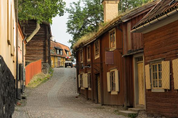 A photo looking up a cobblestone street surrounded by mostly wooden buildings. At the end of the street, we see a tarvern (Krog), which, unlike other buildings has a roof made of shingles. A few buildings to the left of the street seem to be made of stone, but the rest is wooden, with a reddish color (a paint made with left-overs of the ample iron production in the region, which made the wood last much longer). Also at the end of the street ist a small electric car thingie the Skansen employees use to get around (this was past the closing time of the ticket stalls, so the park was almost fully abandoned).
