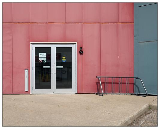 Double doors with white frames set in a wall divided into red and bluish-gray panels, with signs including "Notice Security Cameras in Use" posted on the glass. The doors are flanked on the right by an empty, tipped metal bike rack on a concrete ground.