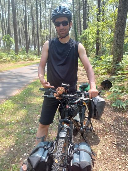 Me standing with my black bike on the side of a cycling path, in a forest. I'm wearing shorts and a sport T-shirt, helmet and sun glasses. My bikes has 4 travel bags, and a plush monkey is holding my bike.