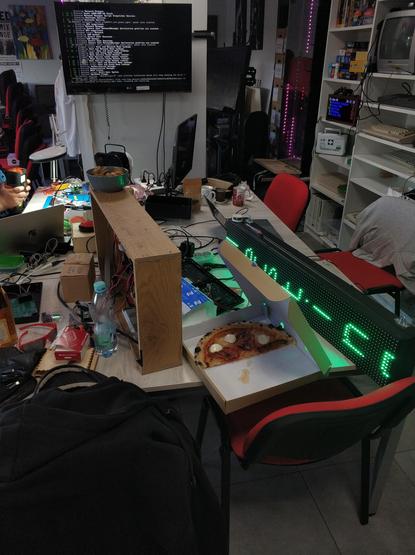 Messy makerspace table full of diy parts, electronic components and devices, screens, etc., and a half-eaten box of pizza. A LED matrix screen laying on its side is showing a green, scrolling text that says "testing". A TV in the background is filled with computer operating system boot messages