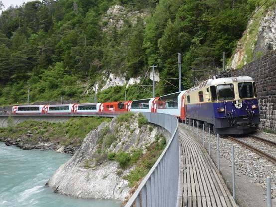 Ein Zug kurvt entlang einer Bahnstrecke in der Nähe eines Flusses, umgeben von grünen Hügeln und felsigen Klippen.