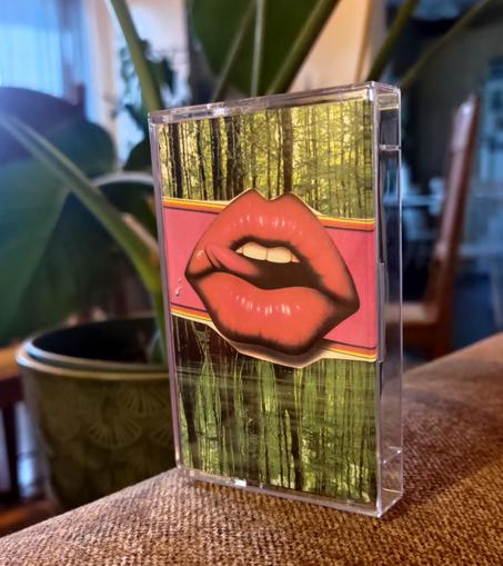 Tape on the back of a couch, with a green plant in the background. 

The cover is a very green image of trees (a forest), with a mostly ping band in the middle, a open mouth in the middle, with big pink lips. The tongue is sticking out, licking the lip. A drop of water/spit is seen on the left side.