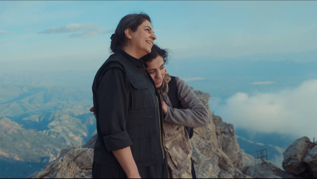 Dos mujeres de mediana edad abrazadas miran al infinito en lo alto de una montaña en medio de una cordillera, las rodean el cielo azul, las nubes blancas y las montañas pardas