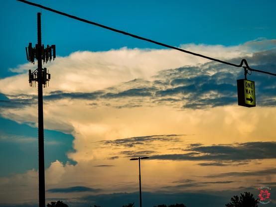 Photo of a sunset with a cloud formation in the far background sky, with a 5G tower and a one-way street post hanging in the foreground