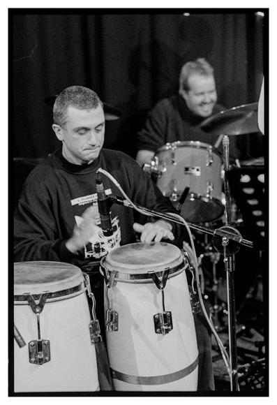 Black and white photograph of a concentrated José J. Cortijo (percussion) in the foreground and a laughing Wolfgang Haffner (drums) in the out-of focus background.