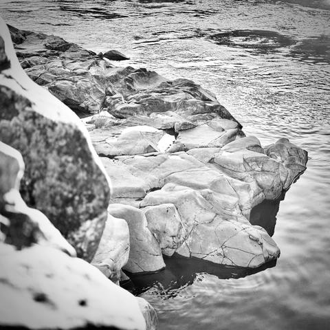 Shallow rocks rise out of the flowing water in the shade of a sunny morning, as taller rocks on frame left of the foreground reflect its rays of light, all in black and white.